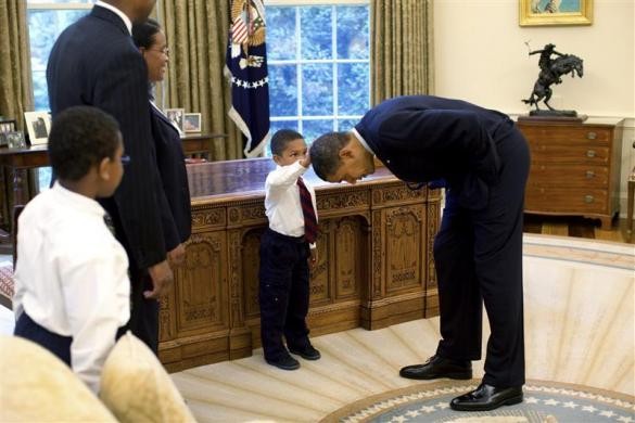 President Barack Obama bends over so that the son of a White House staff member can pat his head during a family visit to the Oval Office, May 8, 2009. Barack Obama