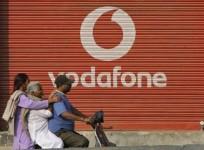 A man and two passengers ride on a scooter past a shop displaying the Vodafone logo on its shutter in Jammu