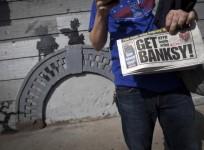 A man holds up a local newspaper in front of an art piece by British street artist Banksy in New York.