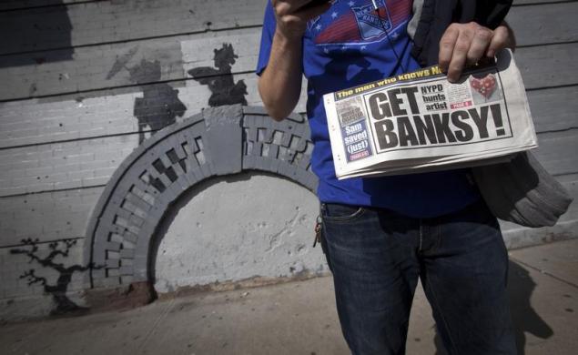A man holds up a local newspaper in front of an art piece by British street artist Banksy in New York.