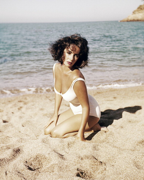 Elizabeth Taylor (1932-2011), British actress, wearing a white swimsuit as she kneels on the sands of a beach in a publicity still issued for the film, 'Suddenly, Last Summer', 1959. The drama, directed by Joseph L Mankiewicz (1909-1993) and adapted from the play by Tennessee Williams (1911-1983), starred Taylor as 'Catherine Holly'. Beach Babes,Beach Babes pics,Beach Babes images,Beach Babes stills,Beach Babes pictures,Beach Babes photos,Beach Babes hot pics,Beach Babes hot images,Beach Babes hot stills,Beach Babes hot pictures